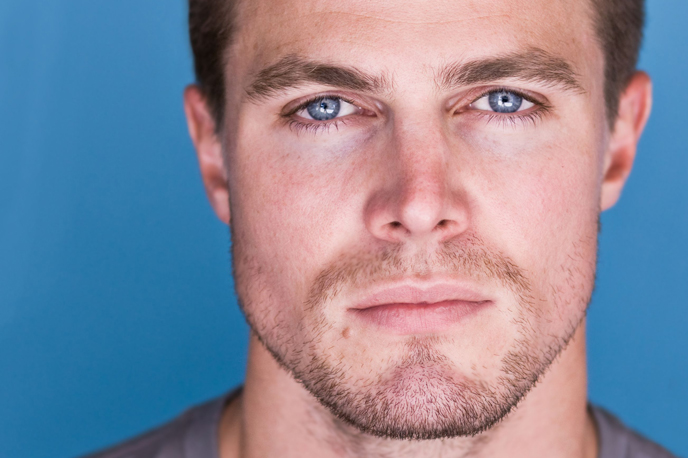 Rosto de um homem jovem com barba por fazer e olhos azuis, em expressão neutra, sobre fundo azul — imagem associada a procedimentos de harmonização facial masculina.